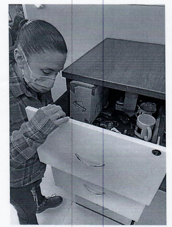 A badly photocopied image of a person wearing a COVID mask searching a filing cabinet. The top drawer has coffee mnugs and other related items.