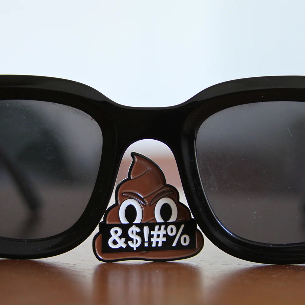 An enamel pin clipped to the nose bridge of black-framed sunglasses resting on a wooden surface. The pin depicts a scowling poop emoji holding a black banner reading "&$!#%". An enamel pin clipped to the nose bridge of black-framed sunglasses resting on a wooden surface. The pin depicts a scowling poop emoji holding a black banner reading "&$!#%".