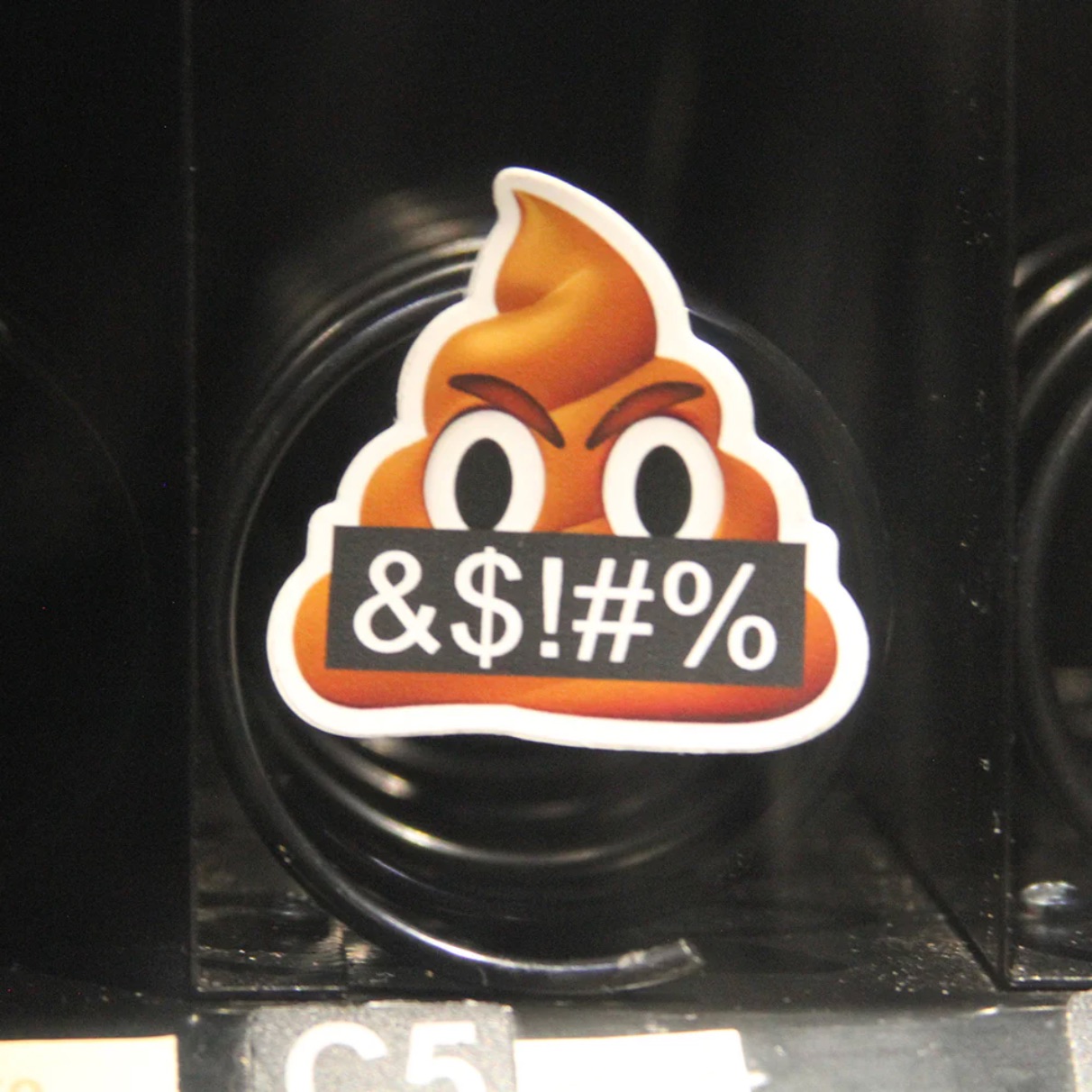 An angry poop emoji sticker affixed to the coiled spring mechanism inside a vending machine. The sticker depicts a scowling poop emoji holding a black sign reading "&$!#%". An angry poop emoji sticker affixed to the coiled spring mechanism inside a vending machine. The sticker depicts a scowling poop emoji holding a black sign reading "&$!#%".